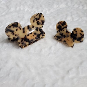 May include: Two tortoise shell hair clips shaped like Mickey Mouse ears. The larger clip is in the foreground and the smaller clip is in the background.