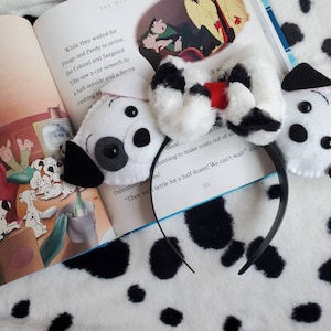 May include: A black headband featuring plush Dalmatian dog ears and a bow. The ears are white with black spots and black details. The bow is white with black spots and a red centre. The headband is placed on a book with Dalmatian illustrations.