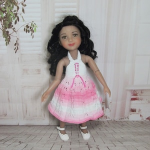 Peut inclure: Une poupée portant une robe en crochet rose et blanche avec un décolleté halter. La poupée a de longs cheveux bouclés foncés et des yeux bruns.