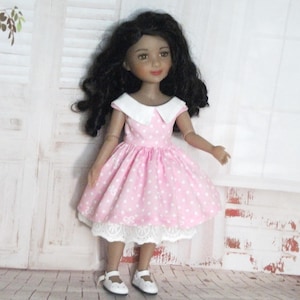 Peut inclure: Poupée vêtue d'une robe rose à pois blancs, col blanc et bordure en dentelle. La poupée a des cheveux noirs et porte des chaussures blanches. Elle se tient sur un sol en briques.