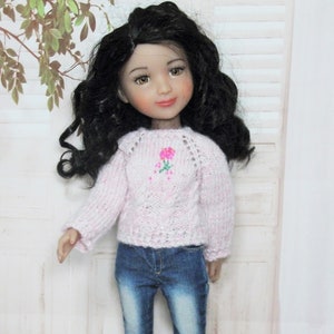 Peut inclure: Un pull rose en tricot avec un motif de fleur sur le devant. Le pull est porté par une poupée avec de longs cheveux noirs bouclés et un jean bleu.