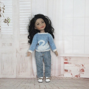 Peut inclure: Une poupée aux cheveux noirs vêtue d'un pull bleu clair avec un motif de cygne blanc, d'un jean bleu et de chaussures blanches. La poupée se tient devant une porte et une fenêtre blanches.