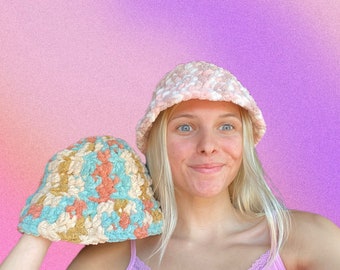 May include: A woman wearing a pink and white crocheted bucket hat and a light pink tank top. She is holding a second crocheted bucket hat in her hand. The hat is a mix of beige, blue, and peach.