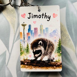 May include: A rectangular keyring featuring a watercolour-style illustration of a raccoon on a log in front of a city skyline with the Space Needle. The name Jimothy is printed in black text at the top, surrounded by small pink hearts and sparkles.