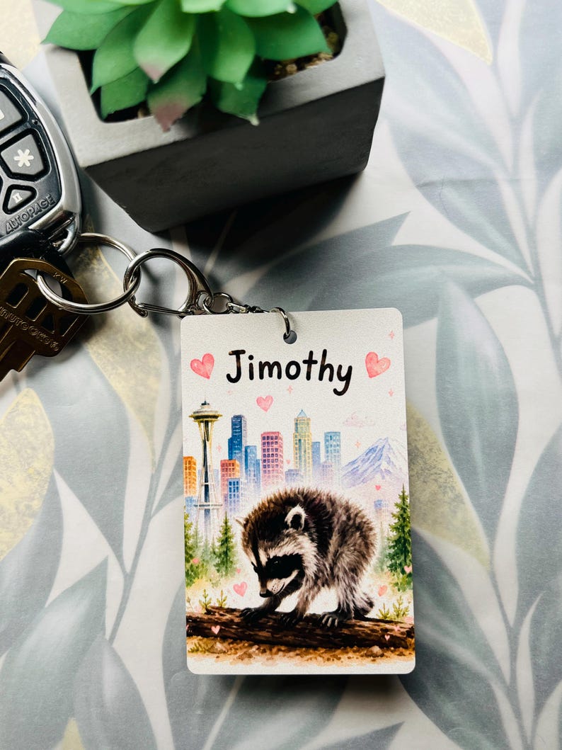 May include: A rectangular keyring featuring a watercolour-style illustration of a raccoon on a log in front of a city skyline with the Space Needle. The name Jimothy is printed in black text at the top, surrounded by small pink hearts and sparkles.