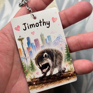 May include: A rectangular keyring featuring a watercolour illustration of a raccoon on a log, a city skyline with the Space Needle, and a mountain. The name Jimothy is printed in black text at the top, surrounded by small pink hearts and sparkles.