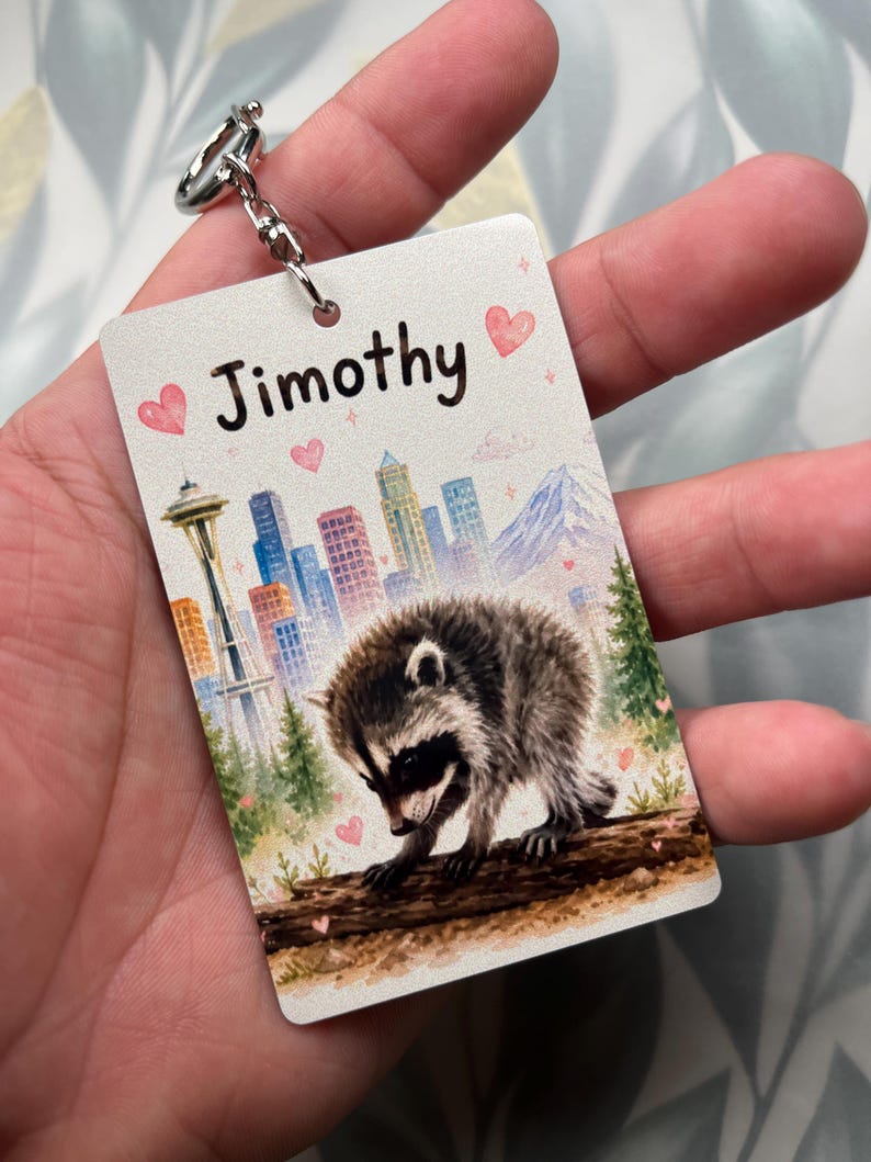 May include: A rectangular keyring featuring a watercolour illustration of a raccoon on a log, a city skyline with the Space Needle, and a mountain. The name Jimothy is printed in black text at the top, surrounded by small pink hearts and sparkles.
