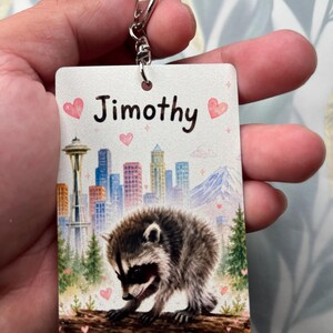 May include: A rectangular keyring featuring a watercolour-style illustration of a raccoon on a log in front of a city skyline with the Space Needle and a mountain. The name Jimothy is printed in black text at the top, accented by pink hearts.