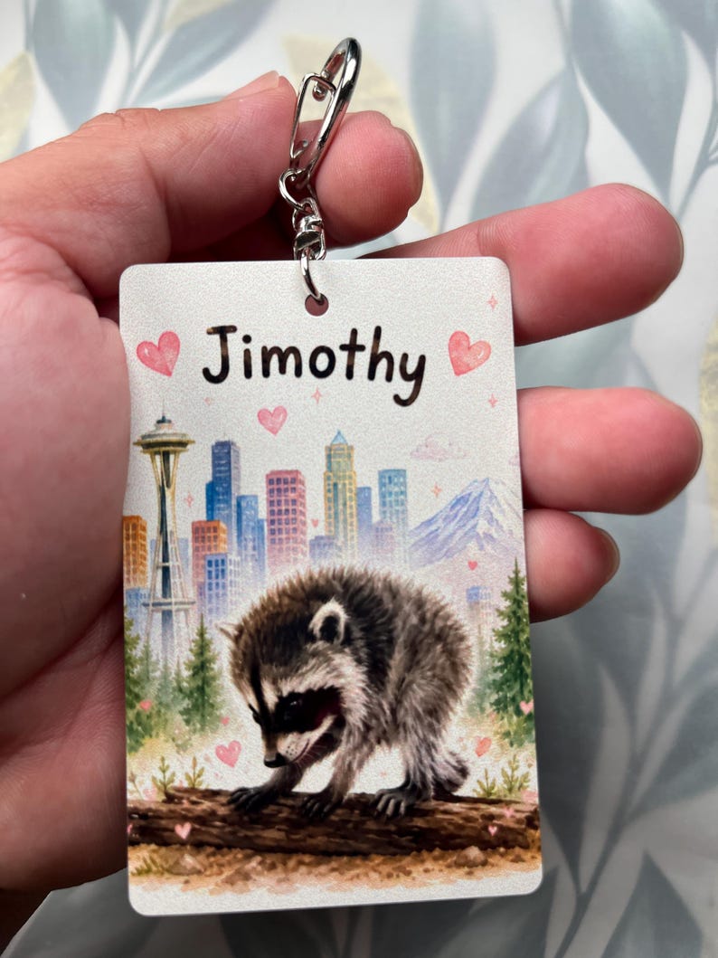 May include: A rectangular keyring featuring a watercolour-style illustration of a raccoon on a log in front of a city skyline with the Space Needle and a mountain. The name Jimothy is printed in black text at the top, accented by pink hearts.