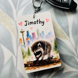 May include: A rectangular keyring featuring a watercolour illustration of a raccoon on a log, a city skyline with the Space Needle, and the name Jimothy printed in black text. The design includes small pink hearts and green pine trees.
