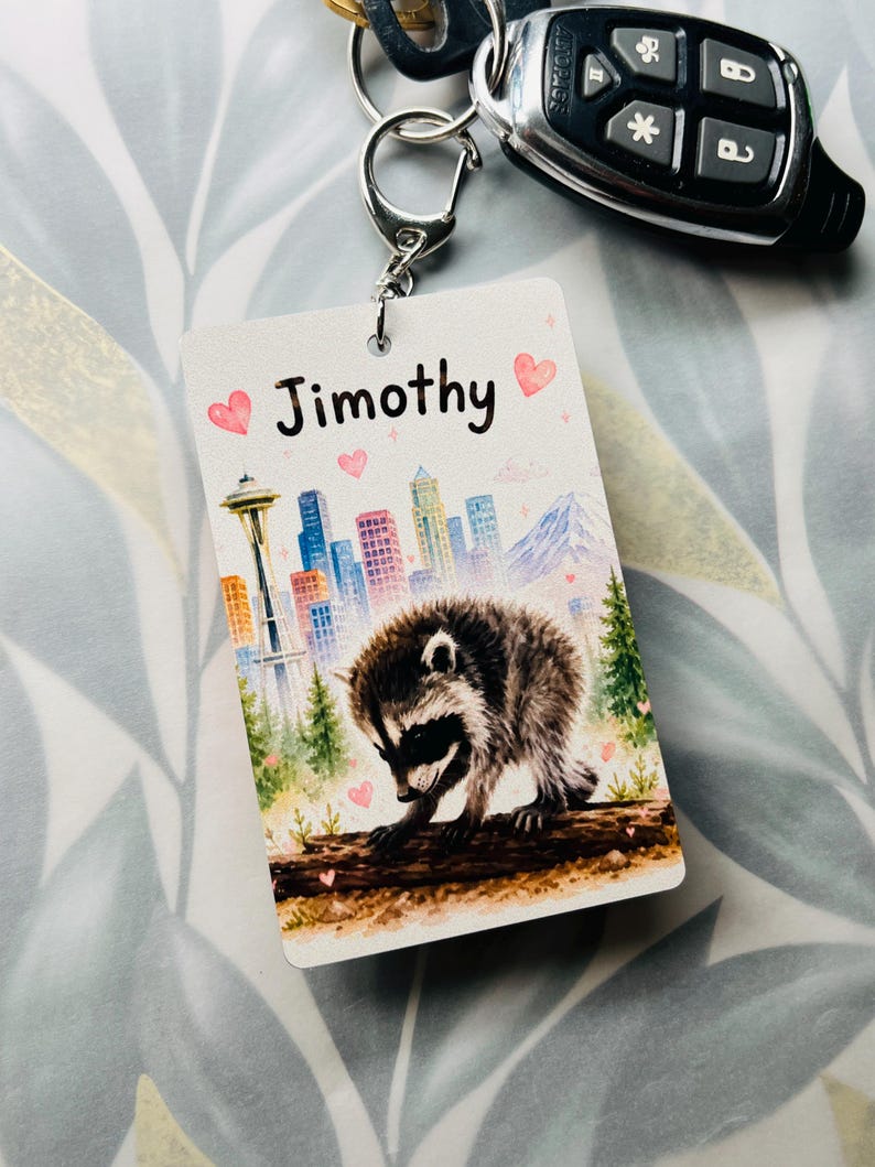 May include: A rectangular keyring featuring a watercolour illustration of a raccoon on a log, a city skyline with the Space Needle, and the name Jimothy printed in black text. The design includes small pink hearts and green pine trees.