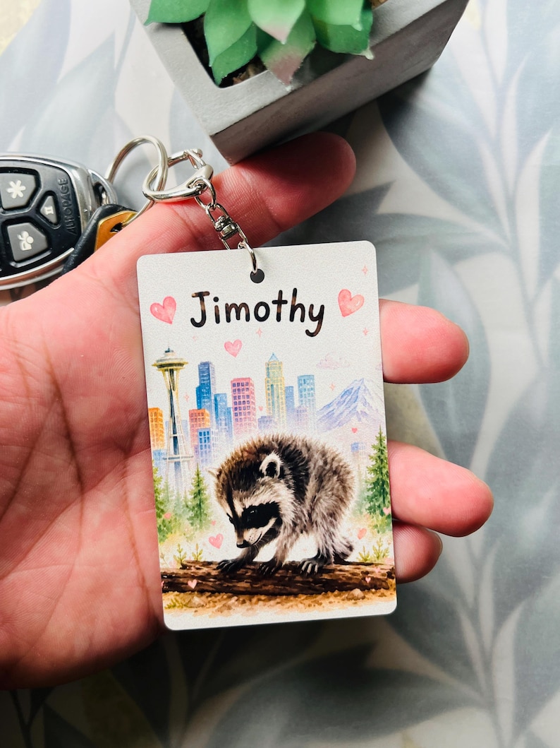 May include: A rectangular keyring featuring a watercolour illustration of a raccoon on a log, a city skyline with the Space Needle, and the name Jimothy printed in black text, accented with small pink hearts on a white background.