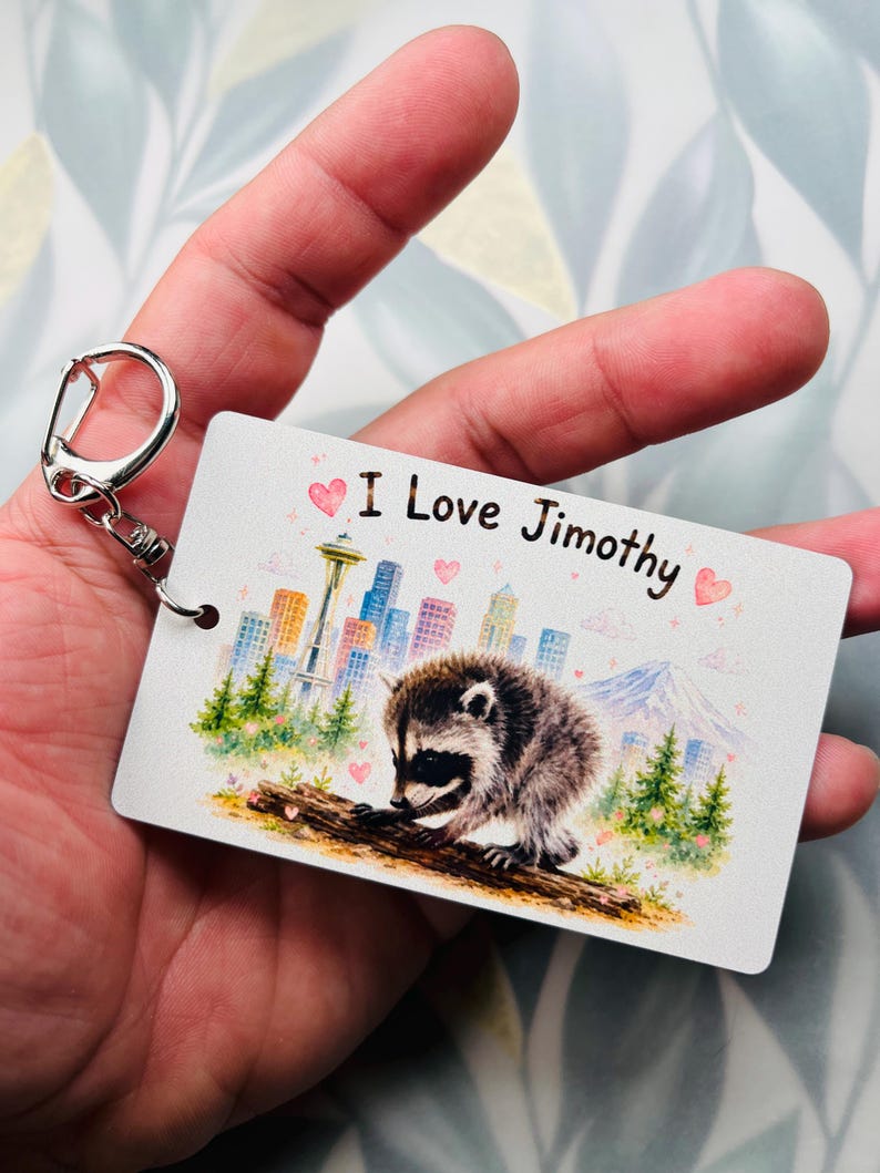 May include: A rectangular white keyring featuring a watercolour illustration of a raccoon on a log, a city skyline with the Space Needle, and a mountain. The text "I Love Jimothy" is printed in black, accompanied by small pink hearts.