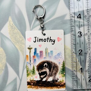 May include: A rectangular keyring featuring a raccoon on a log, a city skyline with the Space Needle, and the name Jimothy. Pink hearts and sparkles decorate the background. A clear ruler shows the keyring is approximately 8.9 cm long.