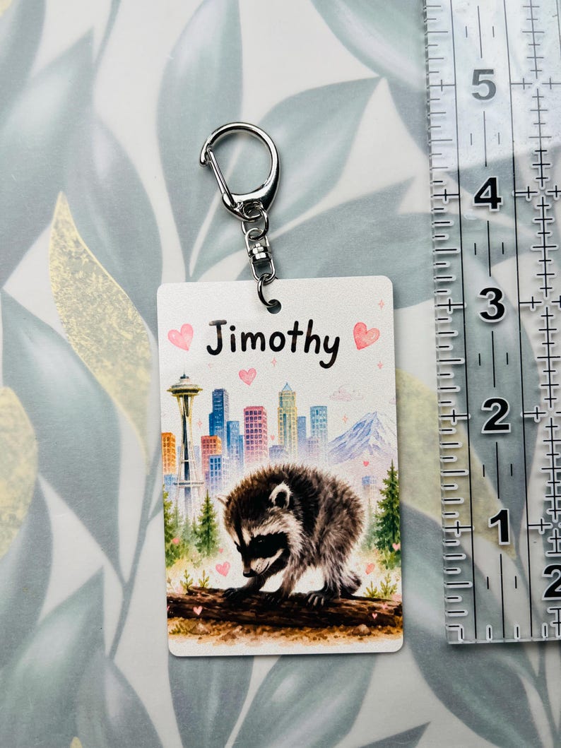 May include: A rectangular keyring featuring a raccoon on a log, a city skyline with the Space Needle, and the name Jimothy. Pink hearts and sparkles decorate the background. A clear ruler shows the keyring is approximately 8.9 cm long.