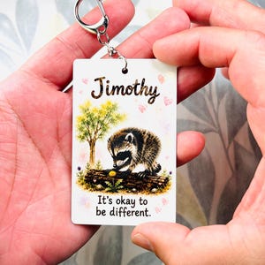 May include: A rectangular white keyring featuring a watercolour illustration of a raccoon on a log near a tree. The text Jimothy is printed at the top, and the phrase It's okay to be different. is printed at the bottom, accented by small pink hearts.