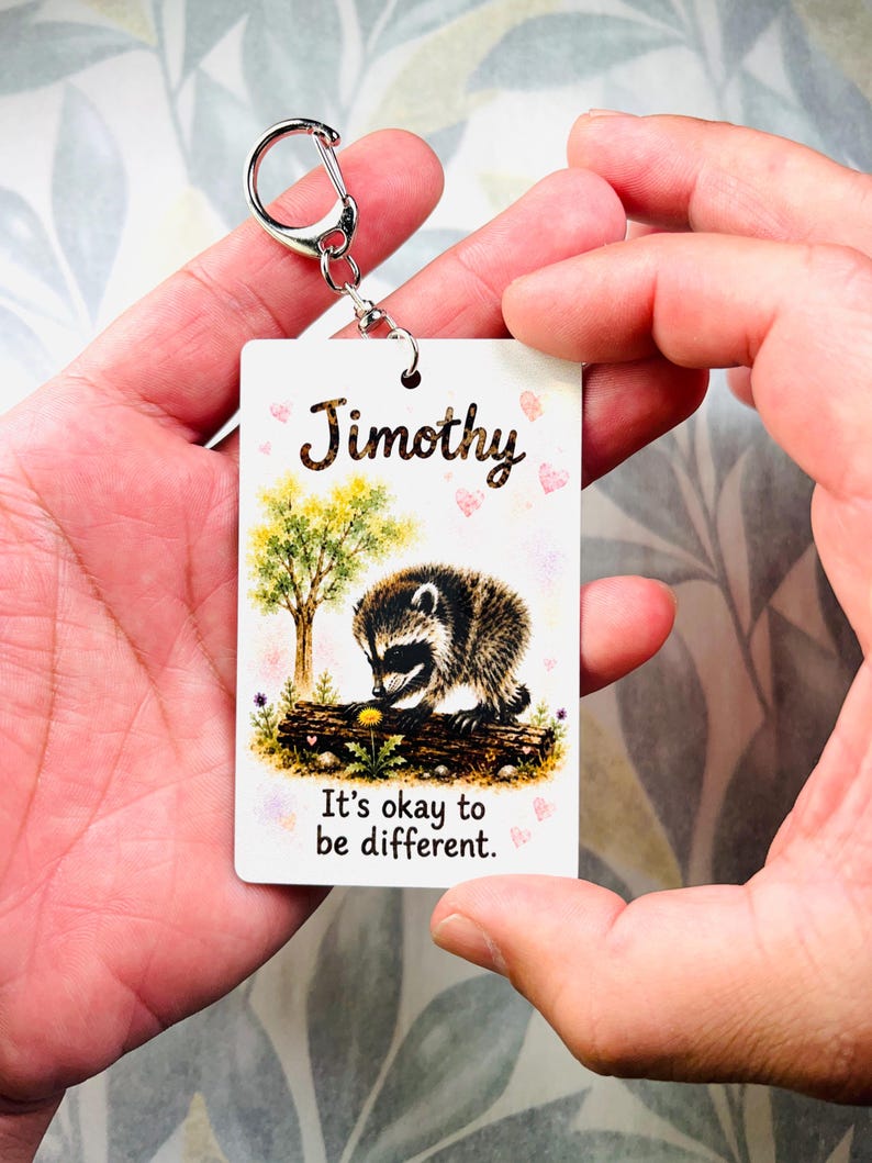 May include: A rectangular white keyring featuring a watercolour illustration of a raccoon on a log near a tree. The text Jimothy is printed at the top, and the phrase It's okay to be different. is printed at the bottom, accented by small pink hearts.
