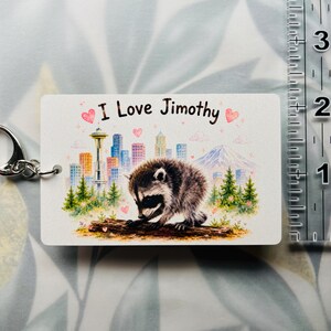May include: A rectangular keyring featuring a raccoon on a log, a city skyline with the Space Needle, and a mountain. The text reads 'I Love Jimothy' with pink hearts. A clear ruler shows the keyring is approximately 6.4 cm wide.
