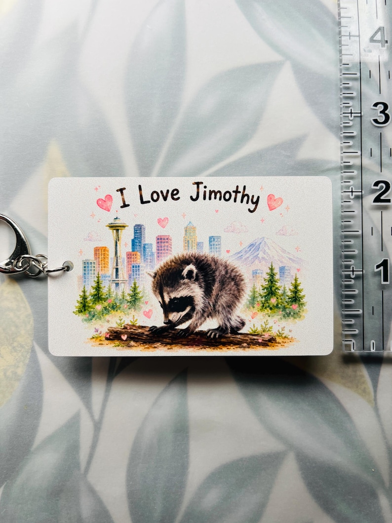 May include: A rectangular keyring featuring a raccoon on a log, a city skyline with the Space Needle, and a mountain. The text reads 'I Love Jimothy' with pink hearts. A clear ruler shows the keyring is approximately 6.4 cm wide.