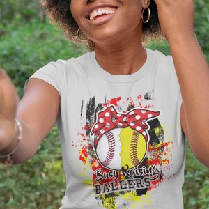 Puede incluir: Camiseta blanca con un dise&ntilde;o de softbol. El softbol est&aacute; dividido en dos, una mitad blanca con costuras rojas y una mitad amarilla, adornado con una diadema roja de lunares. El texto "Busy Raising Ballers" est&aacute; impreso.