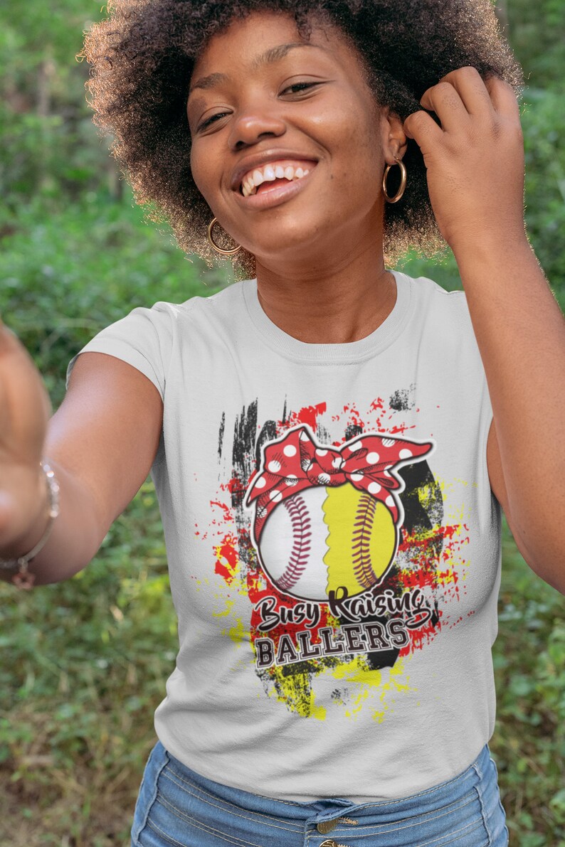 Puede incluir: Camiseta blanca con un dise&ntilde;o de softbol. El softbol est&aacute; dividido en dos, una mitad blanca con costuras rojas y una mitad amarilla, adornado con una diadema roja de lunares. El texto "Busy Raising Ballers" est&aacute; impreso.