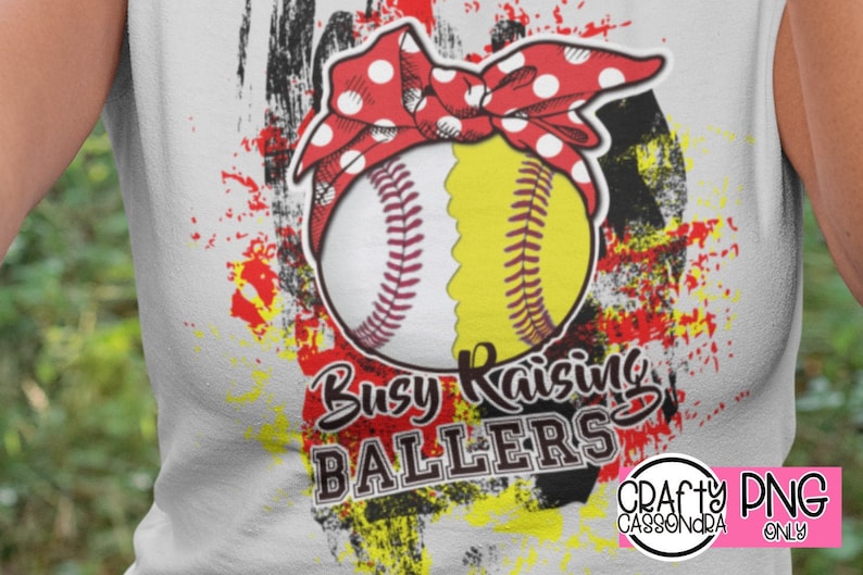 Puede incluir: Camiseta blanca con un dise&ntilde;o gr&aacute;fico de softbol. El softbol est&aacute; dividido entre blanco y amarillo, con una diadema roja con lunares. El texto dice "Busy Raising Ballers". El dise&ntilde;o tiene salpicaduras de pintura roja, negra y amarilla.