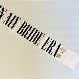 Könnte beinhalten: Weißer Satin-Schärpe mit dem Text "In My Bride Era" in schwarzen Buchstaben. Die Schärpe ist mit goldenen Glitzersternen, einem Diamanten, einer Discokugel und einer Blume verziert.