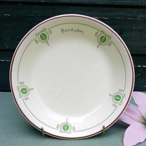 Peut inclure: Une assiette en céramique blanche avec un bord rouge et des détails décoratifs verts et blancs. L'assiette porte l'inscription "Hotel Carlton".
