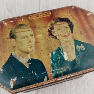 Puede incluir: Una caja de metal rectangular con un retrato vintage de S.M. la Reina Isabel y S.A.R. el Duque de Edimburgo. La tapa muestra a la pareja real con vestimenta formal sobre un fondo cálido y desgastado con arañazos visibles en la superficie.