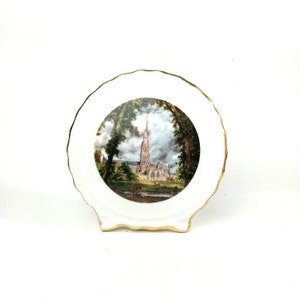 May include: A white ceramic plate with a gold rim features a printed image of a church with a tall spire. The church is surrounded by trees and a grassy area. The plate is on a white background.