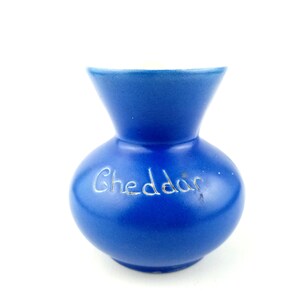 May include: A small, blue ceramic vase with a white rim. The vase has the word "Cheddar" written on the front in white.
