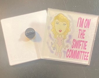 Peut inclure: Un bouton blanc et rose avec une image de dessin animé d'une femme aux cheveux blonds et le texte "I'm on the Swiftie Committee".