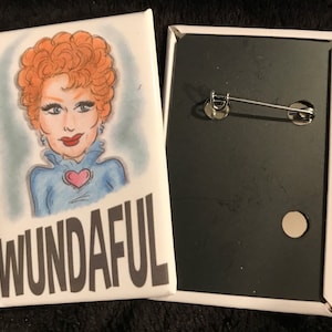 May include: A white button with a cartoon illustration of a woman with red hair wearing a blue shirt with a pink heart on it. The text "WUNDAIFUL" is printed in black at the bottom of the button.