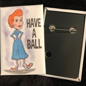 May include: A white button with a cartoon image of a woman with red hair and a blue dress. The text "HAVE A BALL" is printed in black on the button.