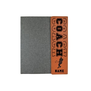 May include: A brown leatherette journal with a gray fabric cover. The front cover has a vertical brown leatherette strip with the word "COACH" in black block letters and a silhouette of a tennis player. The bottom of the strip says "NAME".
