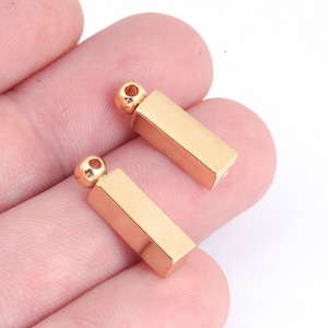 Puede incluir: Dos colgantes de barra de metal de color dorado con un bucle en la parte superior para sujetar a una cadena o cordón.