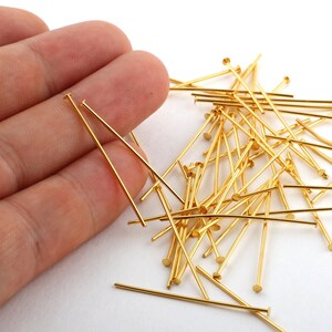 May include: A collection of gold-colored head pins, scattered on a white surface. Each pin has a straight, slender shaft and a small, flat head. The pins are of varying lengths, creating a visually interesting arrangement.