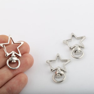 Puede incluir: Tres llaveros de estrella plateados con cierre giratorio y un anillo. Los llaveros están sobre una superficie blanca.