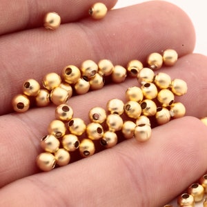 May include: A large quantity of gold-colored metal beads, scattered on a hand. The beads are round and have a smooth, shiny finish.