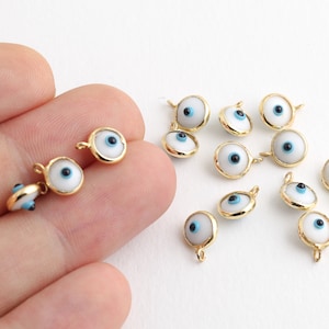 May include: A collection of white and gold evil eye charms with blue centers. The charms are arranged on a white surface.