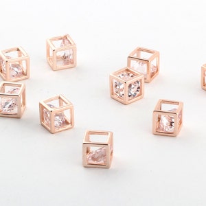 Peut inclure: Neuf breloques en forme de cube en métal doré rose avec des pierres de zircone cubique transparentes à l'intérieur. Les breloques sont disposées sur une surface blanche.