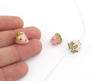 Puede incluir: Cuentas de vidrio rosas con forma de fresa, con puntos rojos y detalles de hojas verdes y amarillas. Una cuenta se sostiene en una mano, mientras que otras dos están ensartadas en un alambre fino de color plateado. Las cuentas están sobre un fondo blanco.