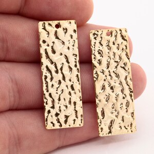 May include: Two gold-colored metal rectangular pendant charms with a textured surface. Each charm has a small hole at the top for attaching a string or chain.