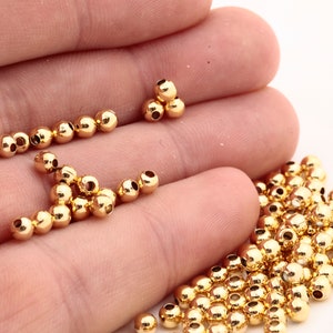 May include: A large quantity of gold-colored metal beads. The beads are round and have a small hole in the center. The beads are scattered on a hand and a white surface.