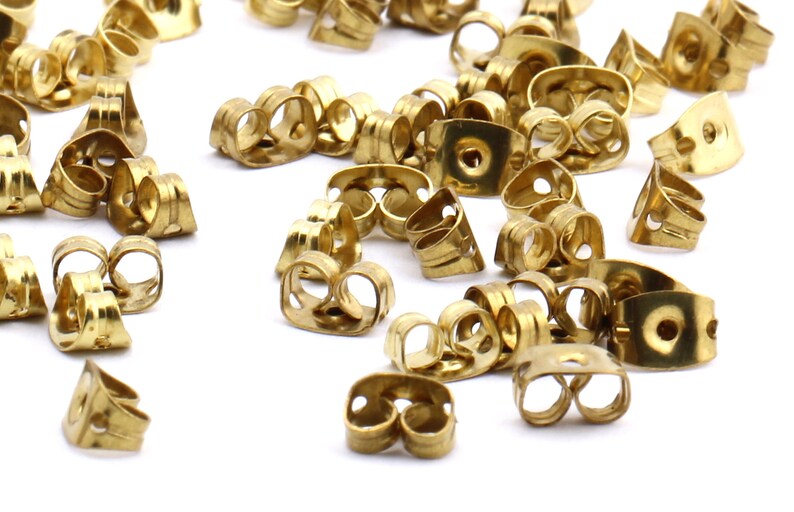May include: A pile of gold-colored earring backs. The backs are made of metal and have a small hole in the center. They are designed to be used with stud earrings.