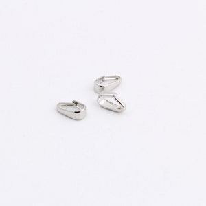 May include: Three small, silver-colored metal jewelry findings, each with a teardrop shape and a hinged top. The findings are scattered on a white background, with a bright, reflective surface.