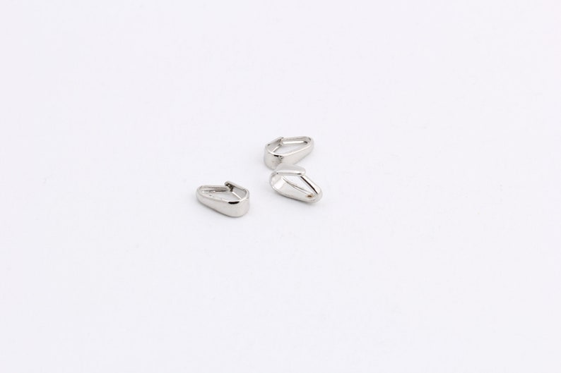 May include: Three small, silver-colored metal jewelry findings, each with a teardrop shape and a hinged top. The findings are scattered on a white background, with a bright, reflective surface.