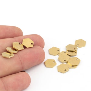 Puede incluir: Un grupo de dijes hexagonales de metal dorado con un pequeño agujero en la parte superior. Los dijes están esparcidos sobre una superficie blanca y se sostienen en una mano.