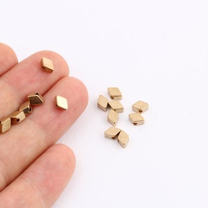 May include: A group of small, gold-colored, diamond-shaped beads with holes in the center. The beads are scattered on a white surface.
