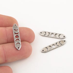 May include: Three silver metal charms shaped like a long rectangle with a crescent moon design cut out. The charms are approximately 5 cm long.